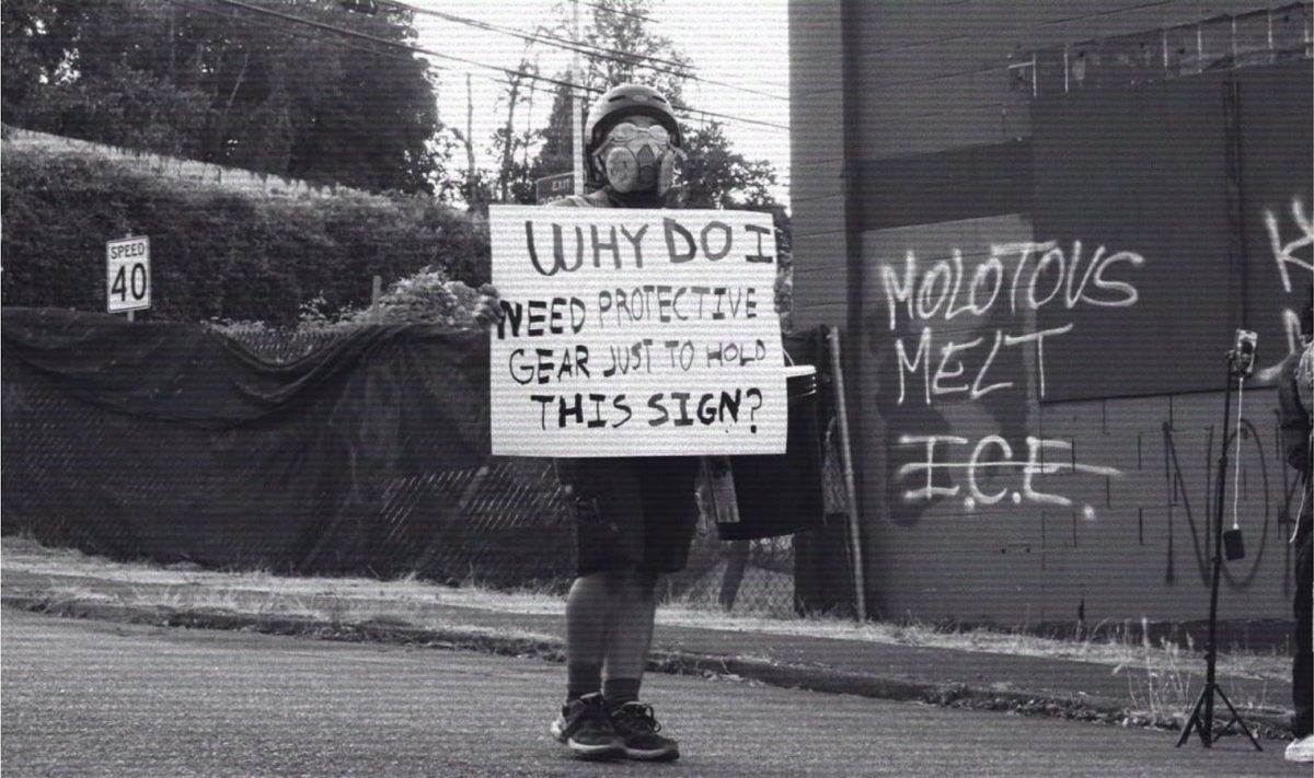 Protester outside the Portland ICE facility with a sign saying “Why do I need protective gear just to hold this sign?” In the background, graffiti reads “Molotovs melt ICE” Molotovs are an improvised incendiary fire weapon used typically by revolutionaries and during riots. 