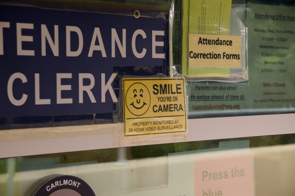 A "Smile, You're on Camera" decal sits beside the attendance clerk window on the side of the administration building, one of the few visible notices that students are being recorded. Its language did not match the wording mandated by the district's own Board Policy 3515.
